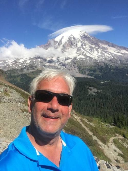Picture of a man with white hair and sunglasses in front of a mountain. 